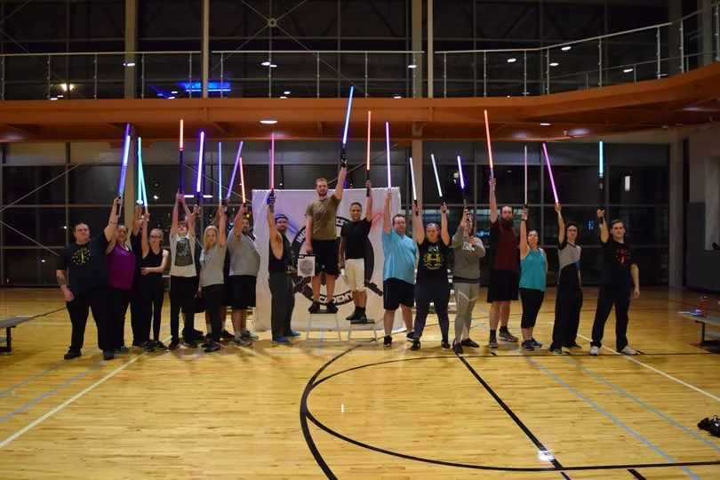A group of Academy participants in a gym, holding illuminated sabers up together.